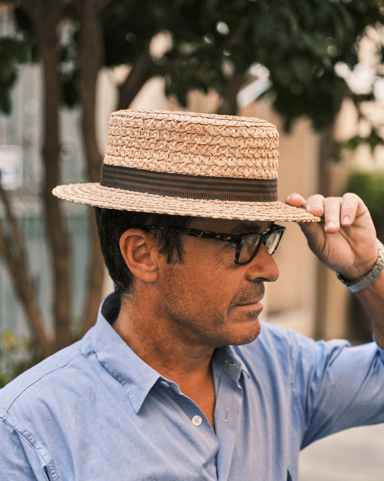 Man wearing a straw hat with black band and light blue shirt, adjusting his hat brim outdoors.