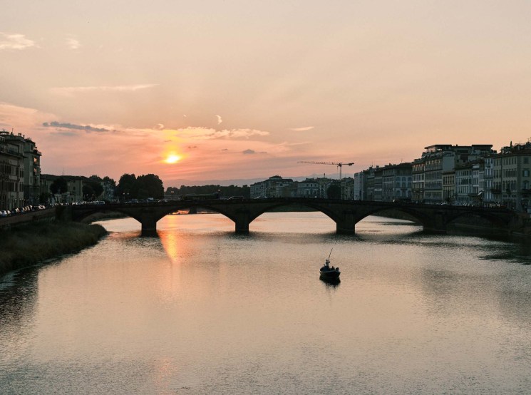 Sunset over the Arno River in Florence with Ponte Vecchio and historic buildings reflected in the water.