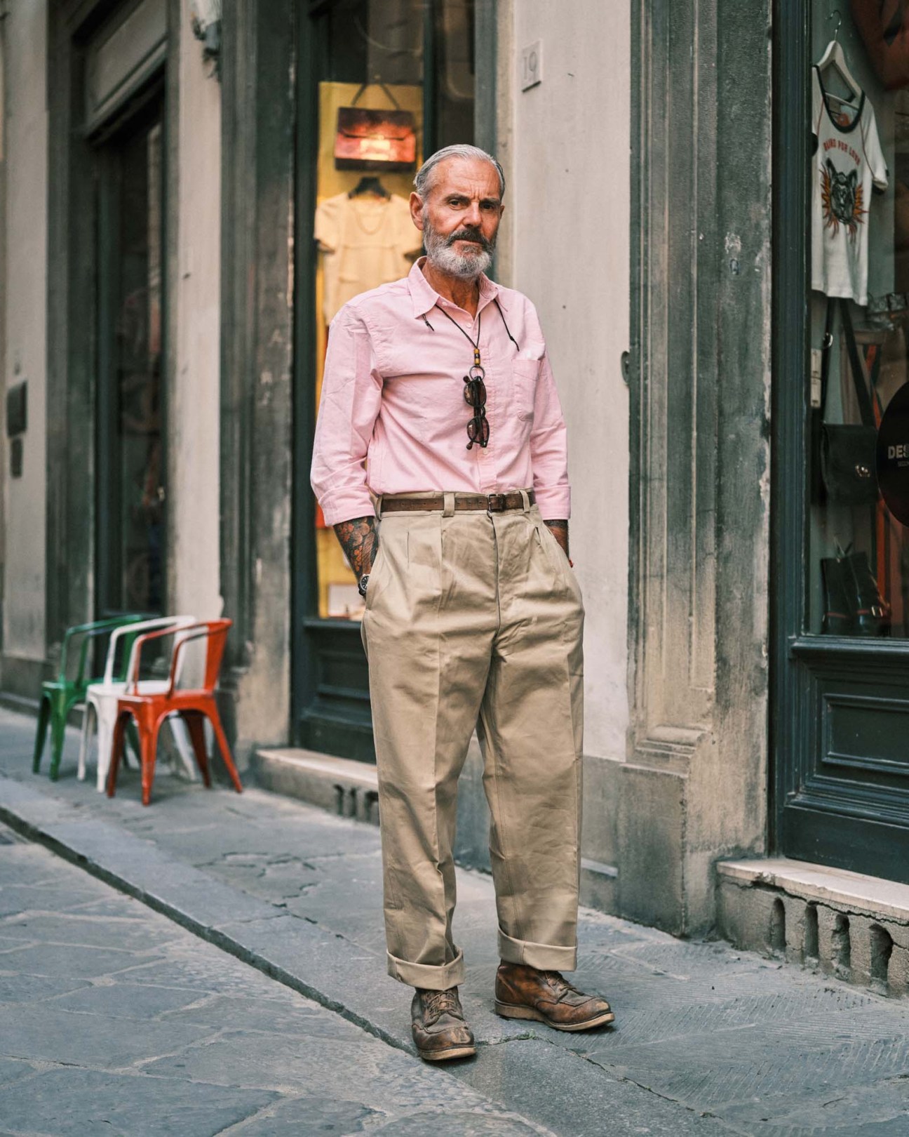 Man in pink shirt and beige chinos standing on a Florence street with colorful chairs and storefronts visible behind him.