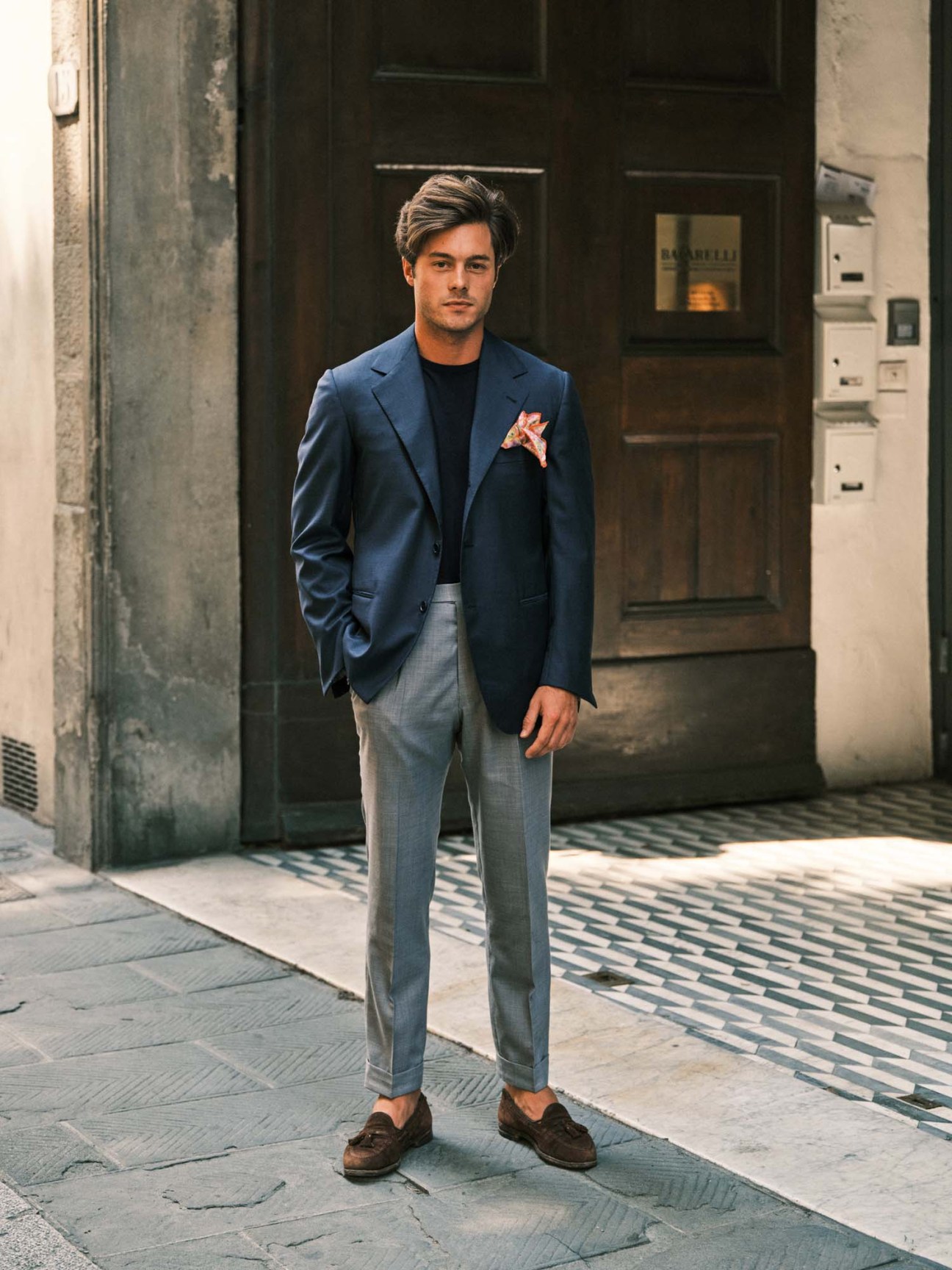 Man in blue blazer, black shirt and grey trousers with coral pocket square stands in Florence doorway.