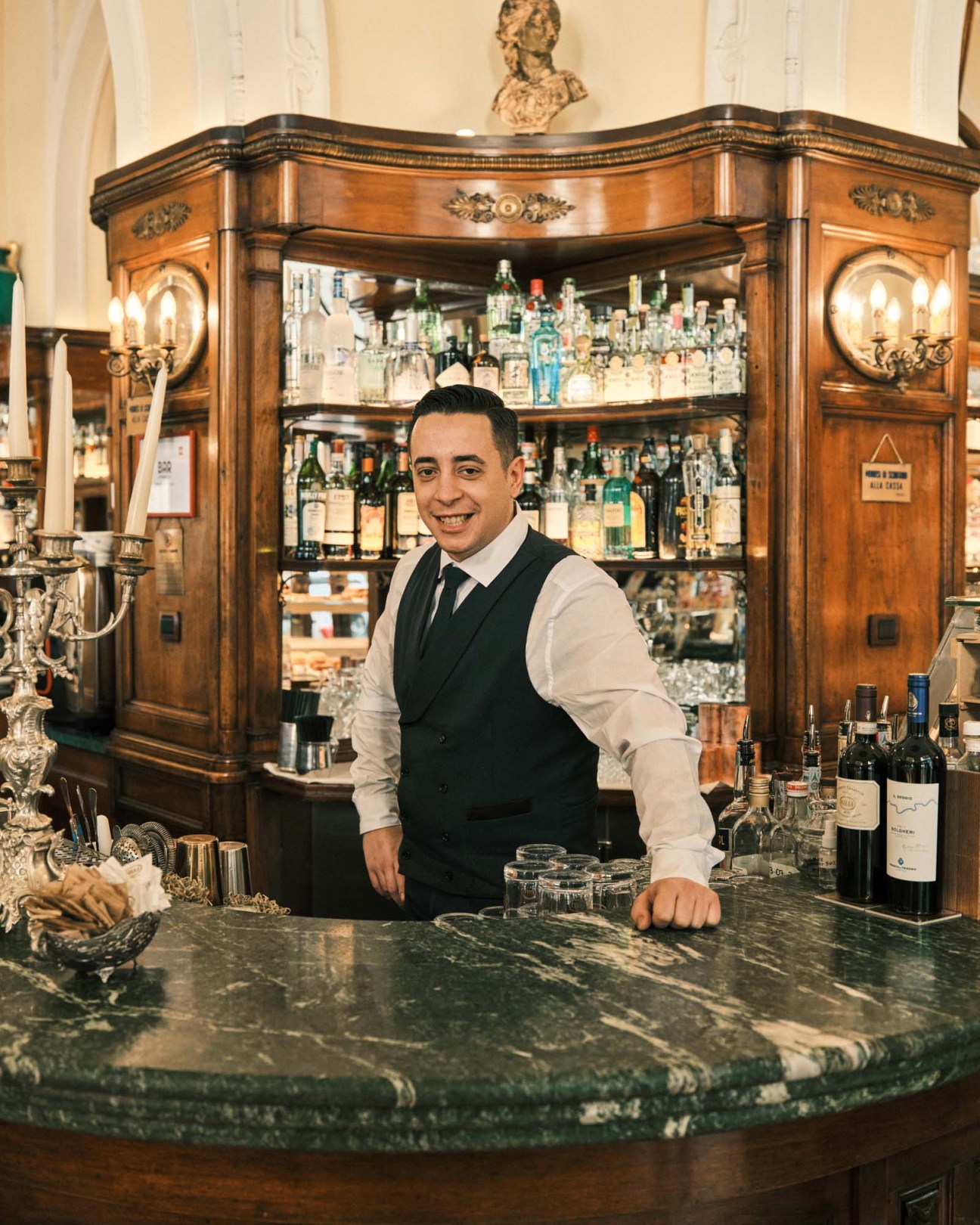 Bartender standing at wooden bar counter with spirit bottles displayed in ornate cabinet behind him.