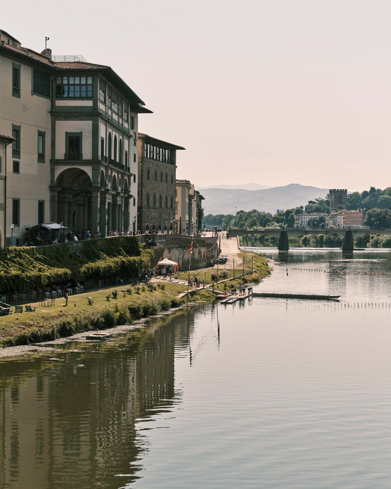 Riverside view of Renaissance palaces and buildings along the Arno River in Florence with cypress trees and hills in the distance.