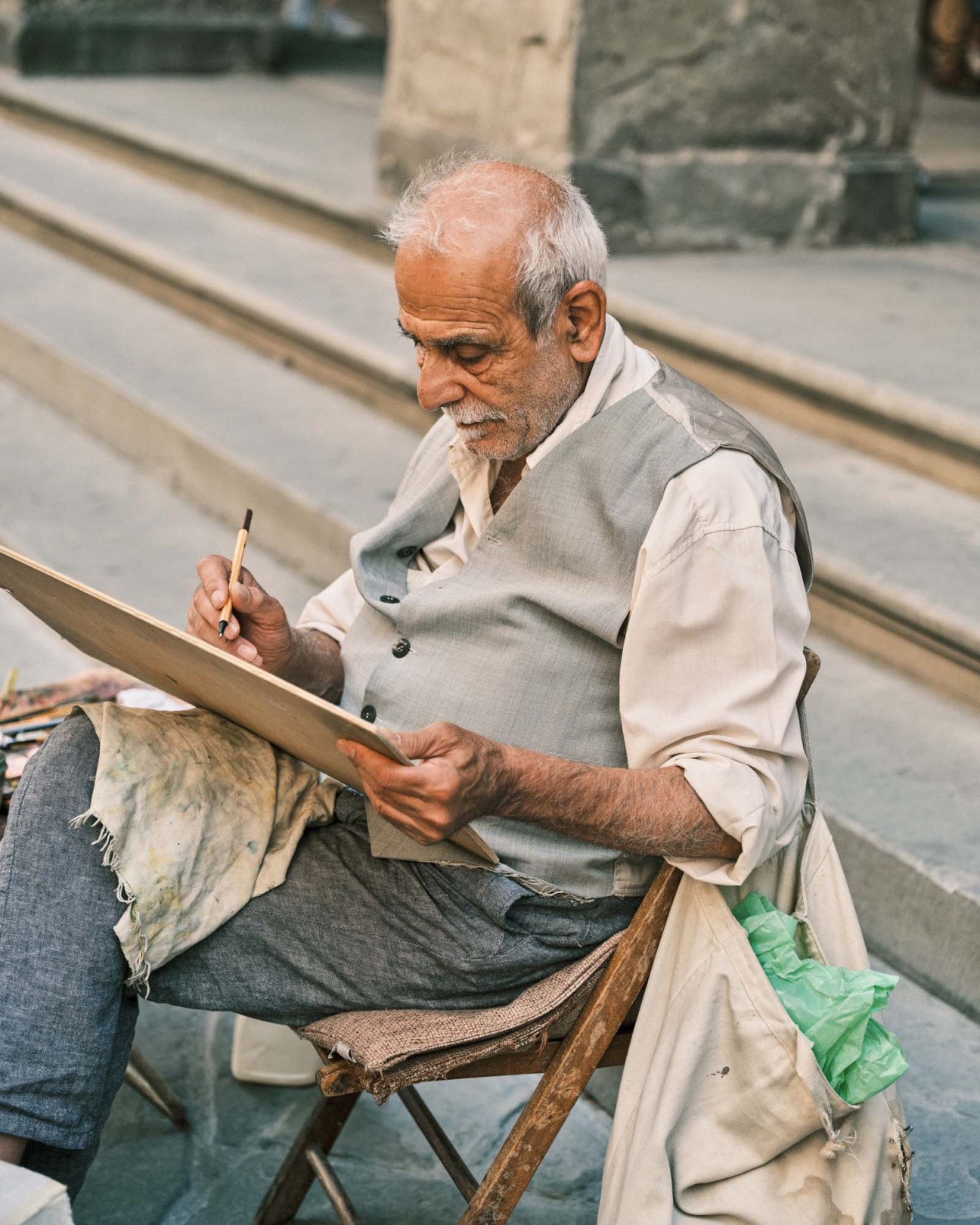 Elderly man in vest and shirt painting on canvas while seated outdoors in Florence.