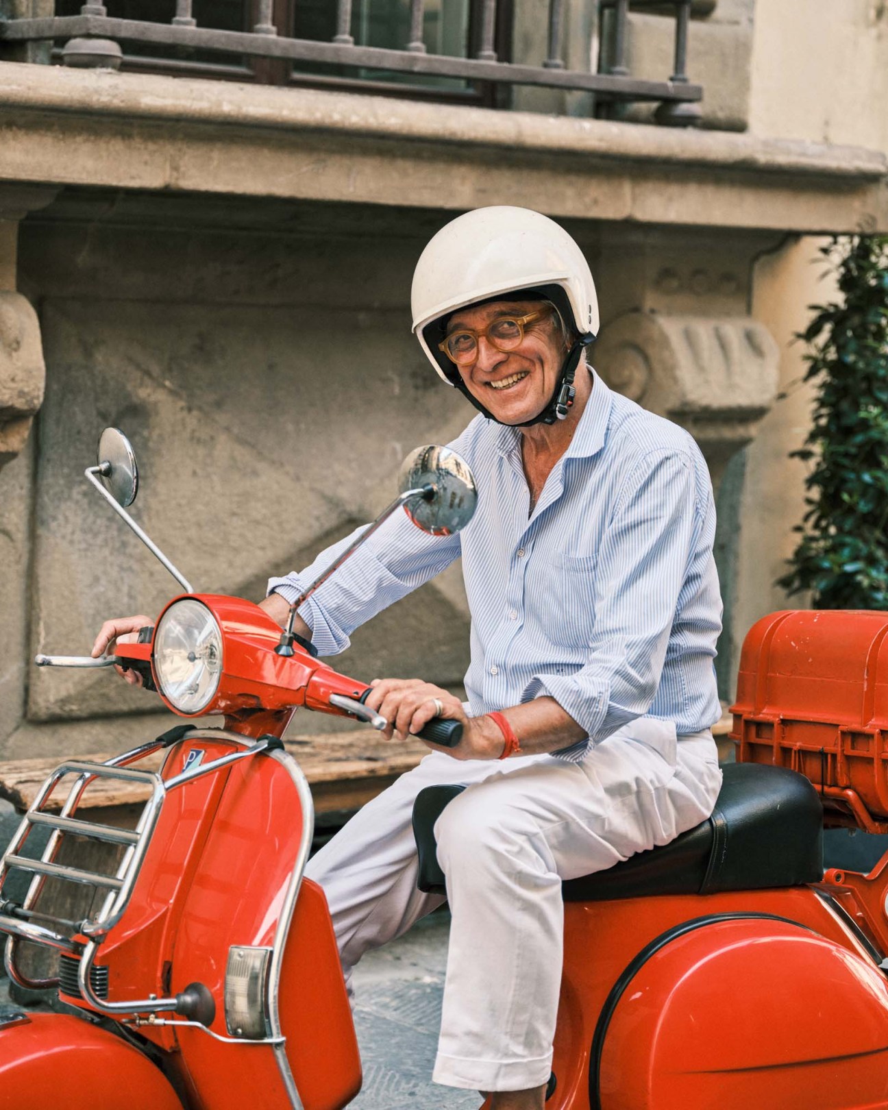 Architect Piero Funis sits on a red Vespa scooter wearing a white helmet, light blue shirt and white trousers, smiling at the camera in front of a stone building.