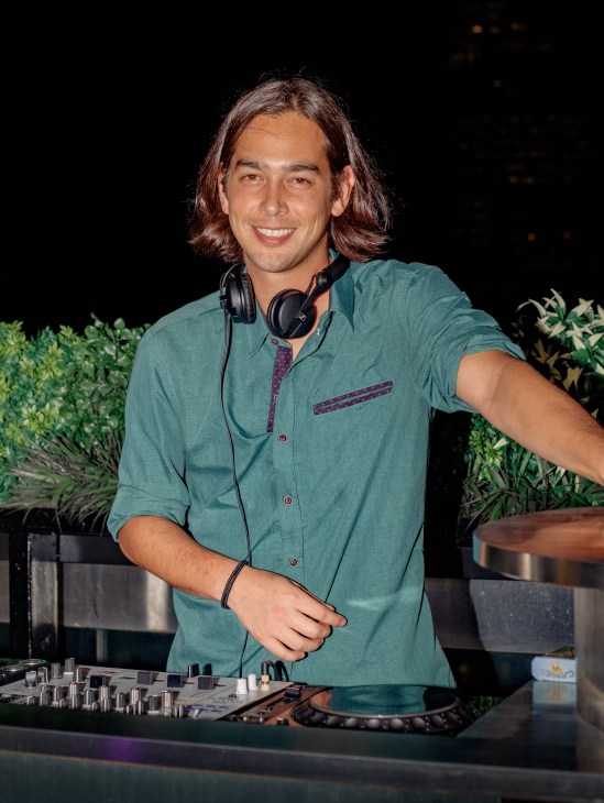 A DJ wearing headphones and a teal shirt smiles while operating a mixing console at night.