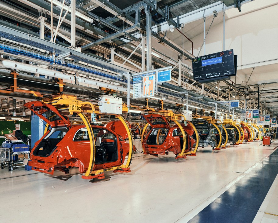 Red Fiat 500e electric cars suspended on yellow overhead assembly line tracks in manufacturing facility.