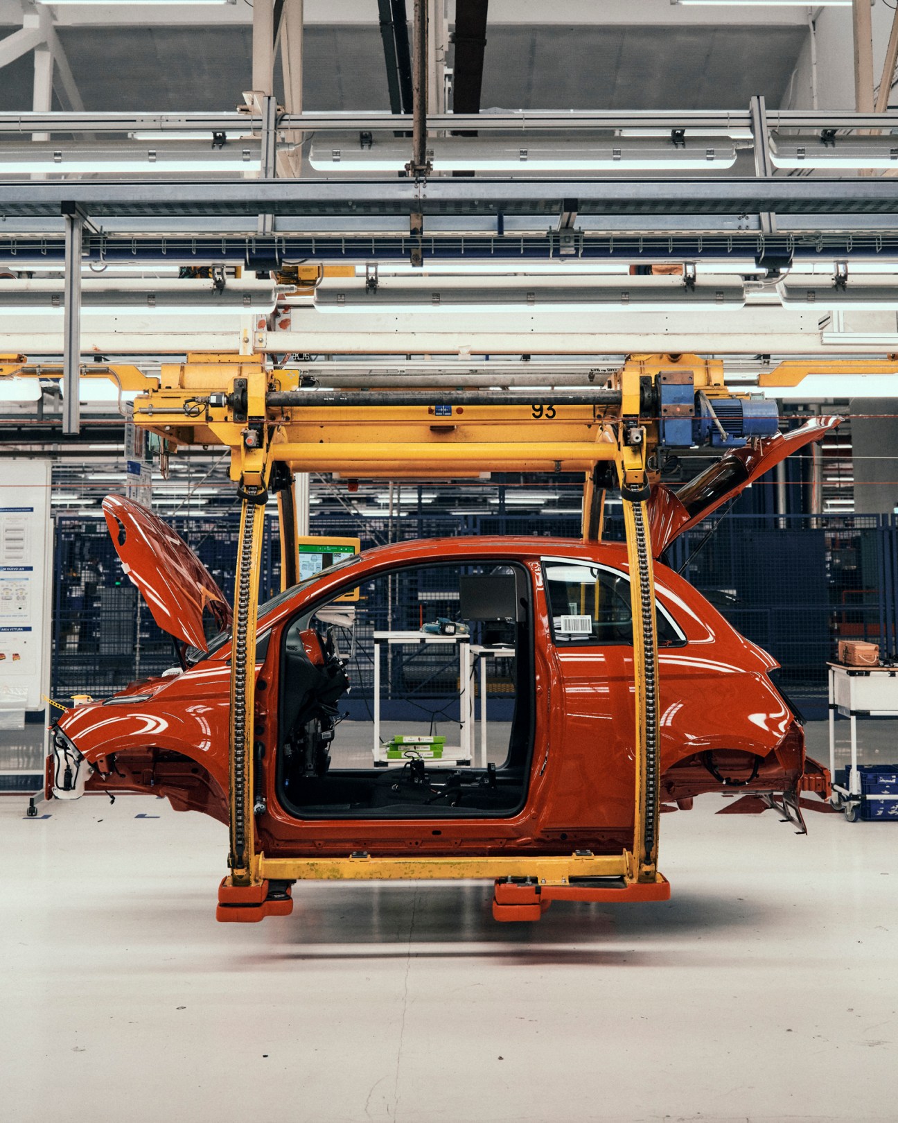 Red Fiat 500e electric car suspended in yellow cradle on assembly line with overhead tracks and robotic arms visible in factory.