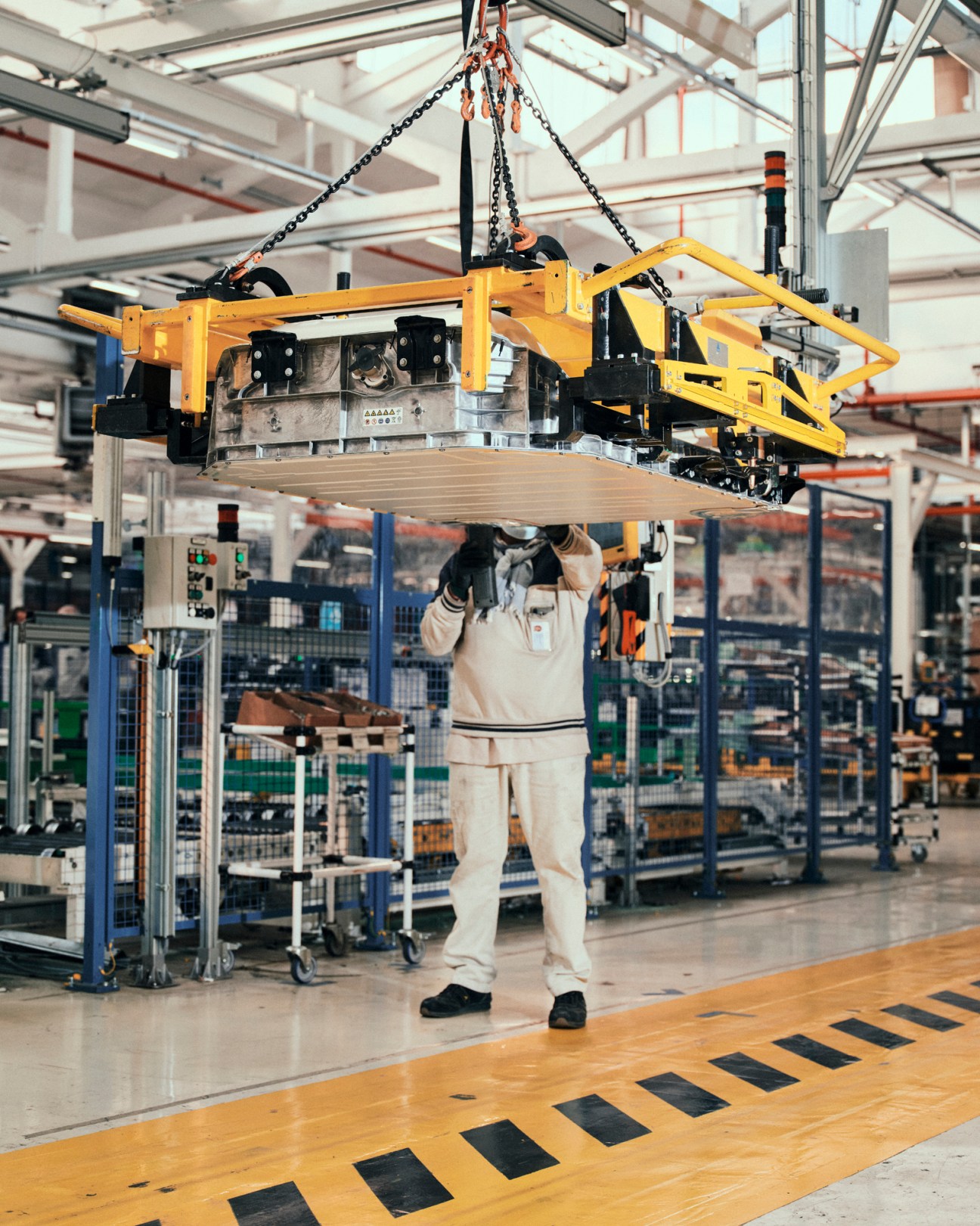 Worker in white uniform assembles Fiat 500e electric car suspended from yellow overhead cradle on production line.