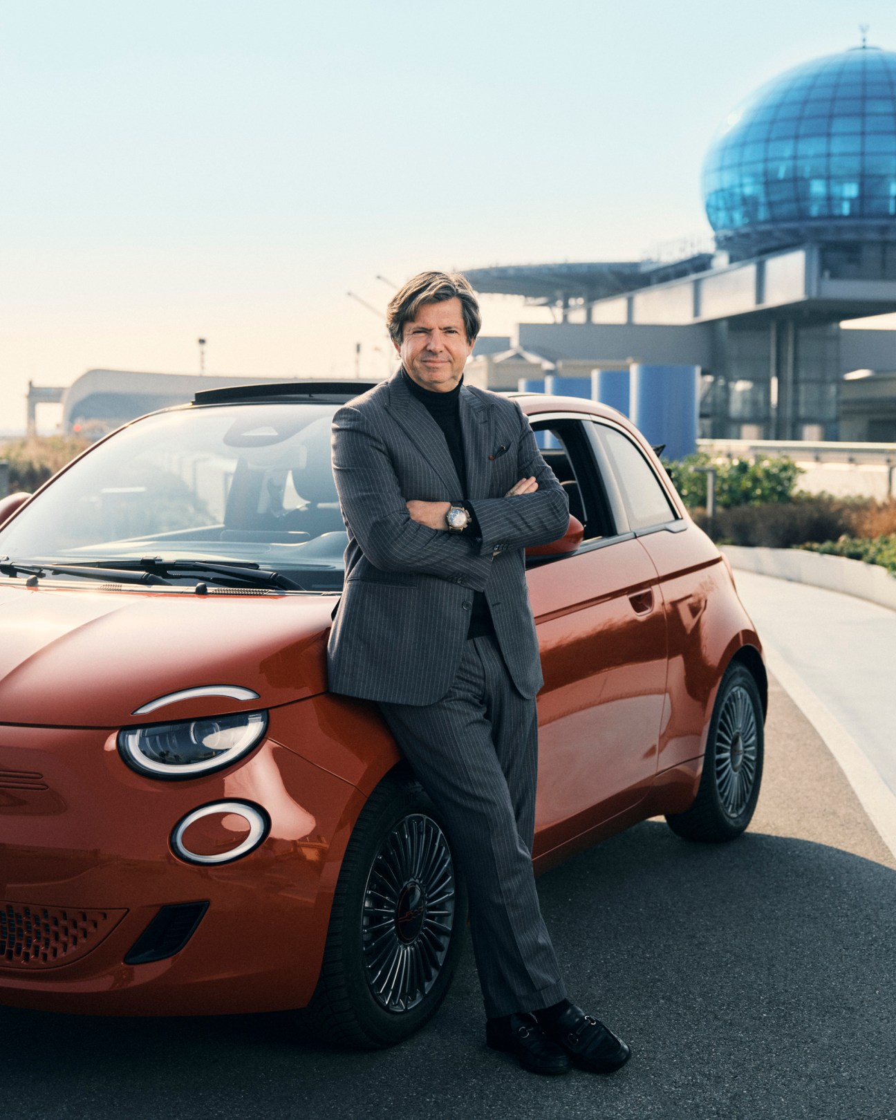 Fiat CEO Olivier François stands with arms crossed next to a terracotta-colored Fiat 500e electric car, with a modern glass dome structure visible in the background.