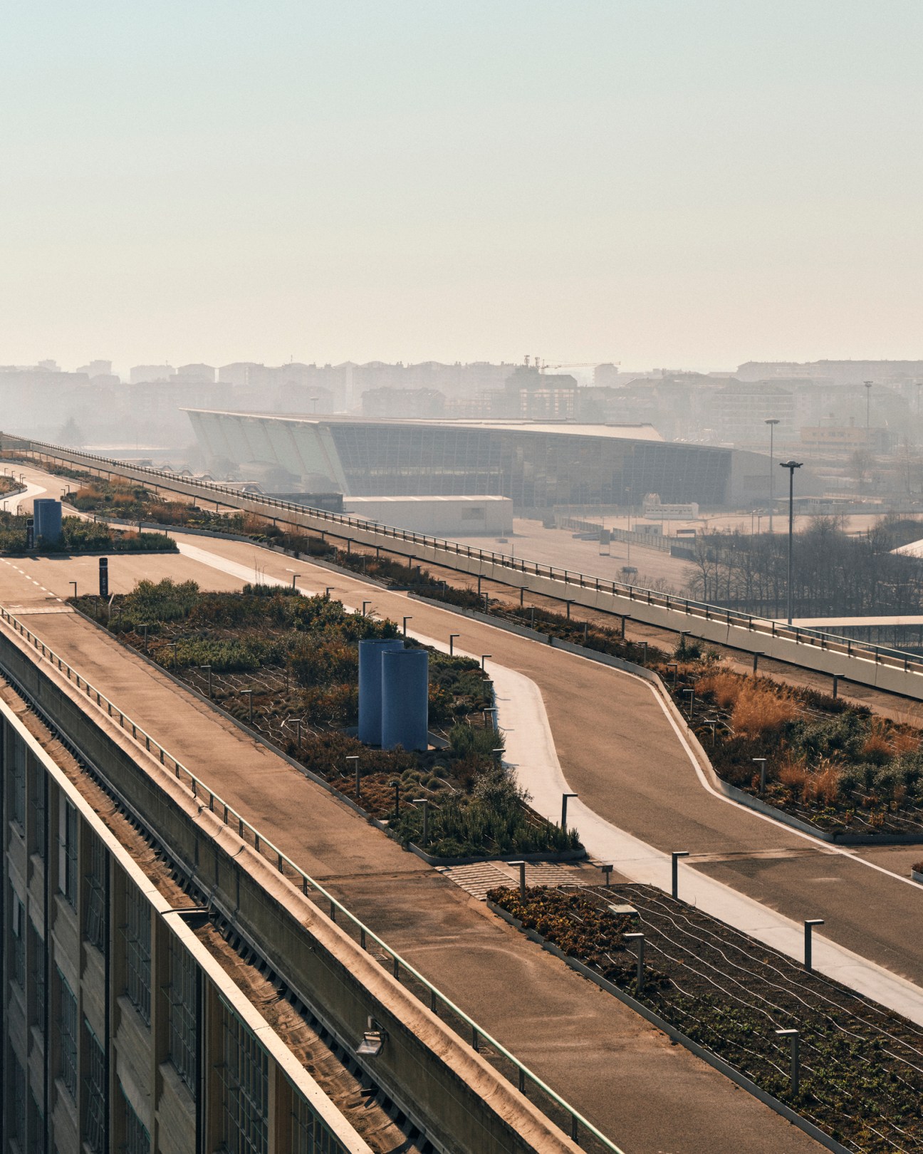 Aerial view of Turin's industrial landscape with the Lingotto building's distinctive angular architecture and rooftop test track visible in the hazy distance.