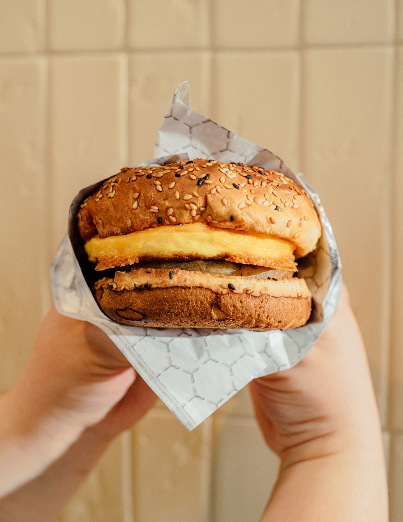 Hands holding a wrapped sausage, egg and cheese sandwich with sesame seed bun