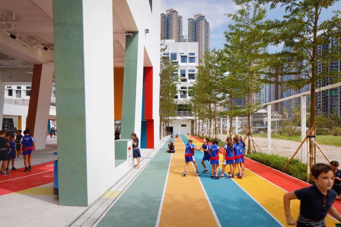 Children playing on a brightly colored running track beneath elevated school building with colorful supporting columns in Hong Kong.