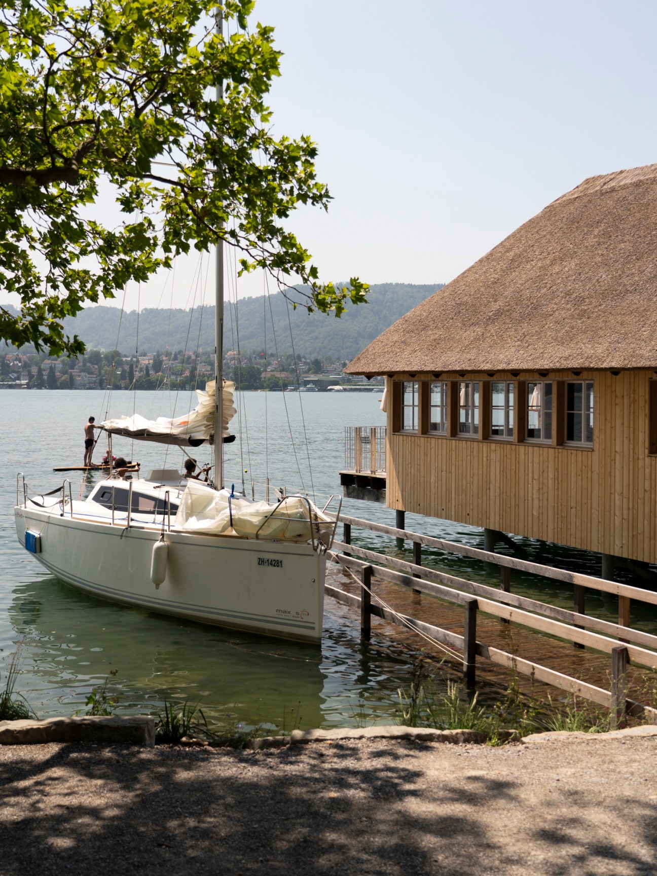 White sailboat moored at wooden pier beside traditional thatched-roof restaurant building on Lake Zürich