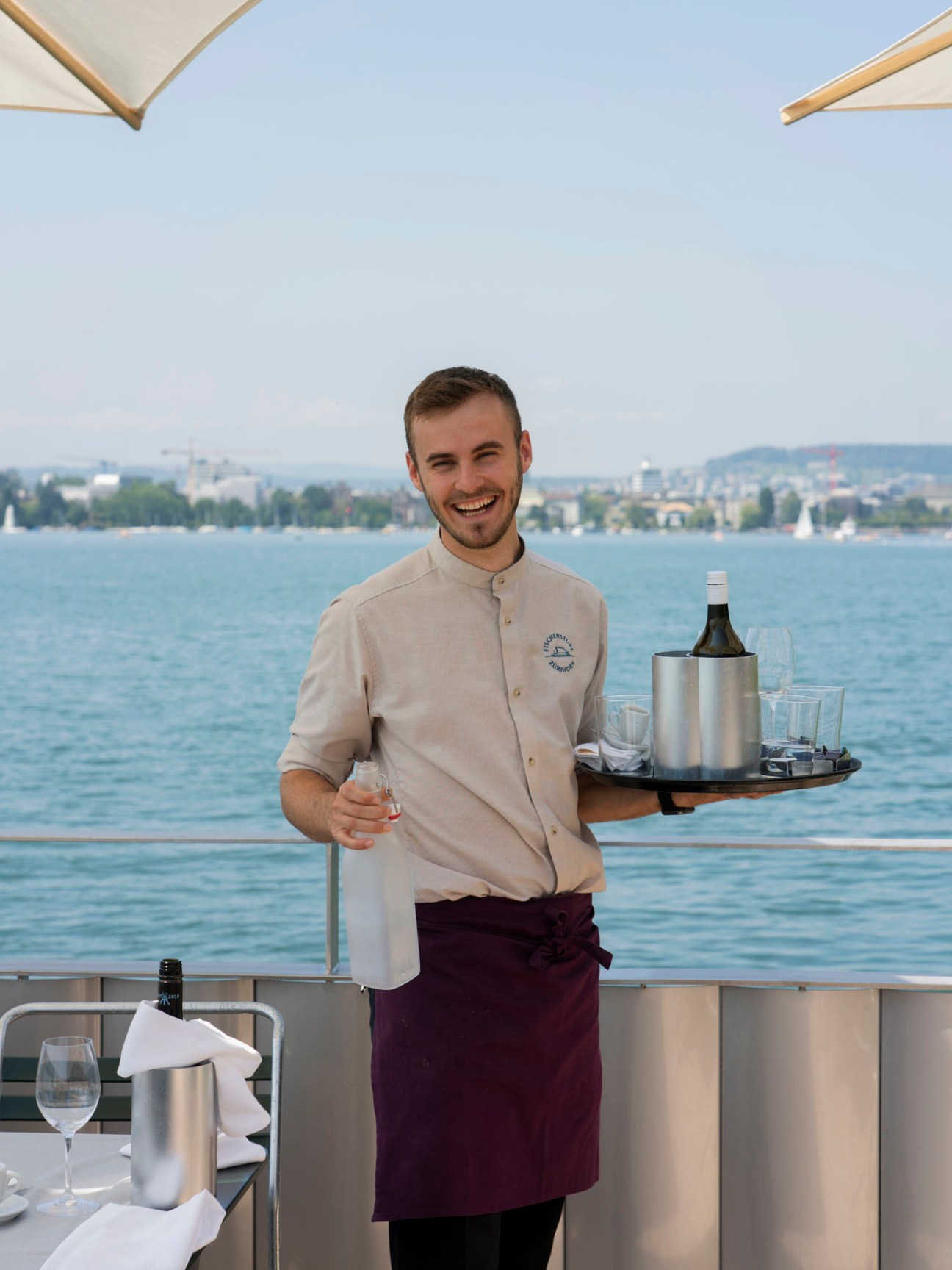 Smiling waiter in beige uniform holding serving tray with wine bottle and glasses on lakeside restaurant terrace