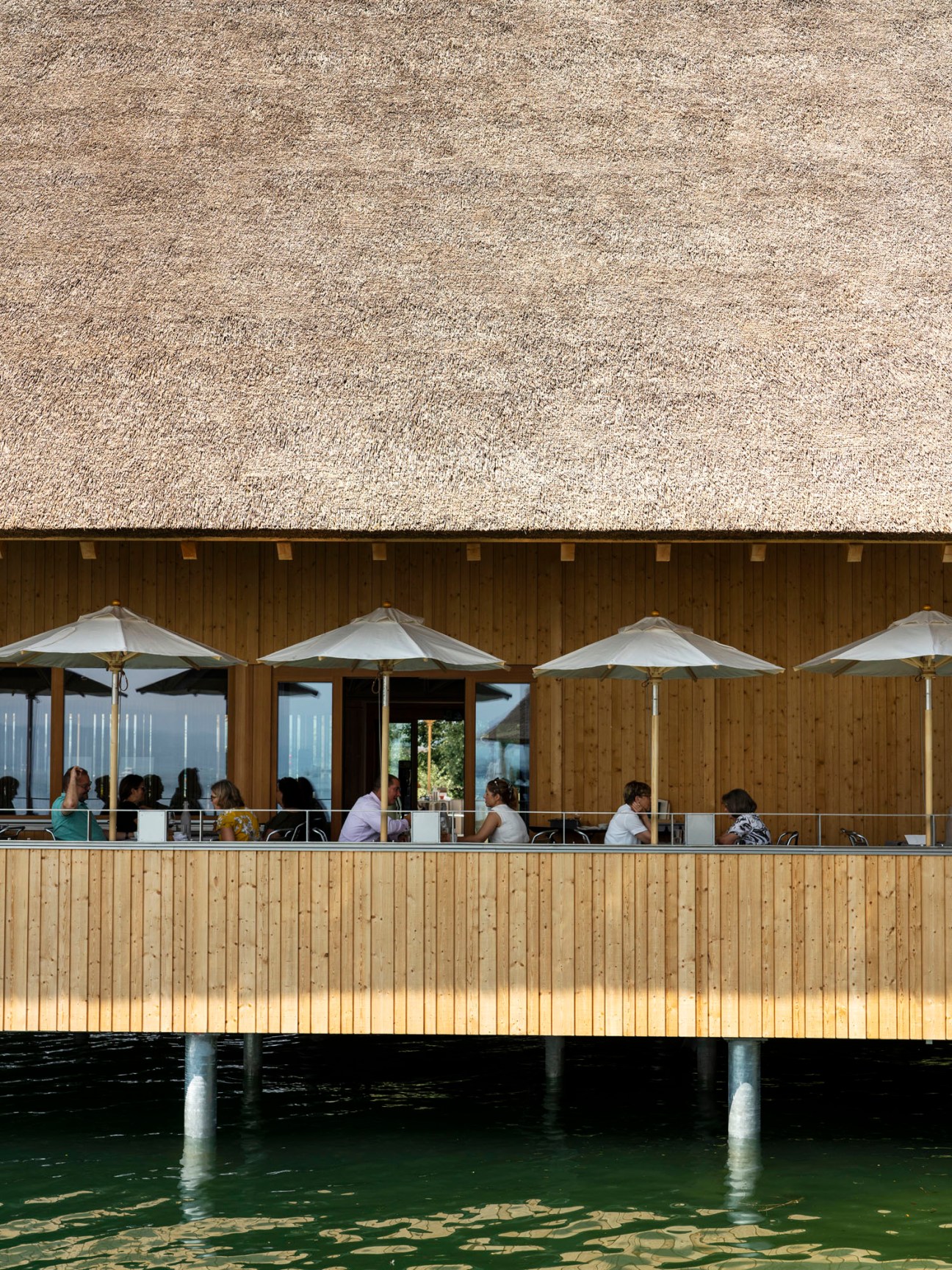 Diners seated at outdoor terrace of wooden restaurant building with thatched roof overlooking water