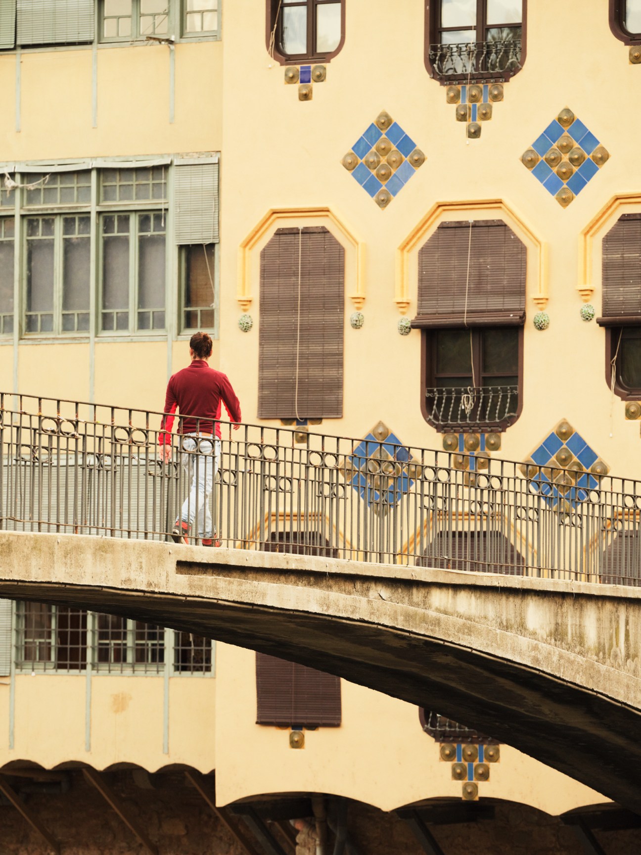 Person in red shirt on concrete bridge with metal railing overlooking yellow building facade with blue tile decorations in Girona, Spain.