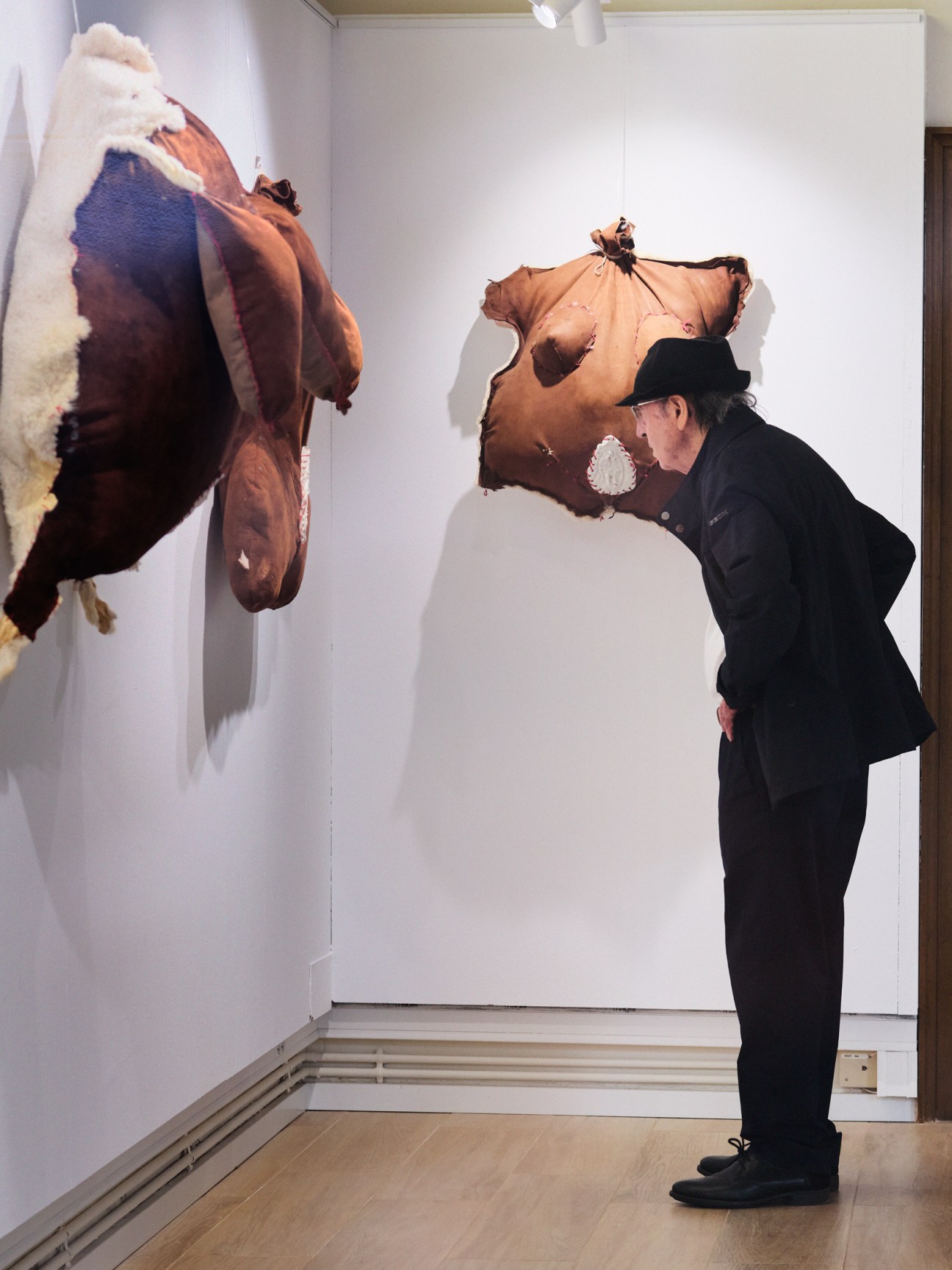 Person in black suit and hat examining abstract brown and white textile artwork hanging on white gallery wall