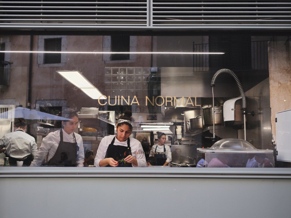 Chefs working in the kitchen of Cuina Normal restaurant with gold lettering visible through the window.