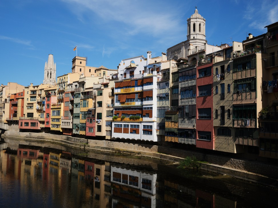 Colorful residential buildings reflected in calm water along Girona's riverfront in Spain.