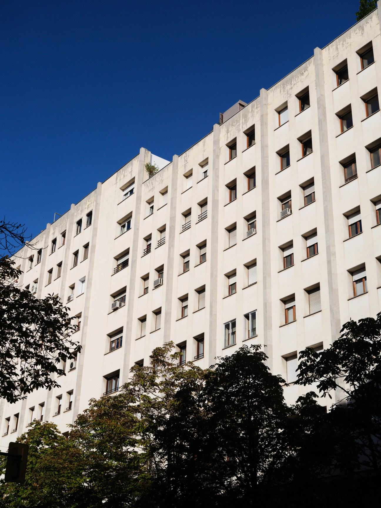 White modernist apartment building with regular window grid against clear blue sky and green foliage below.