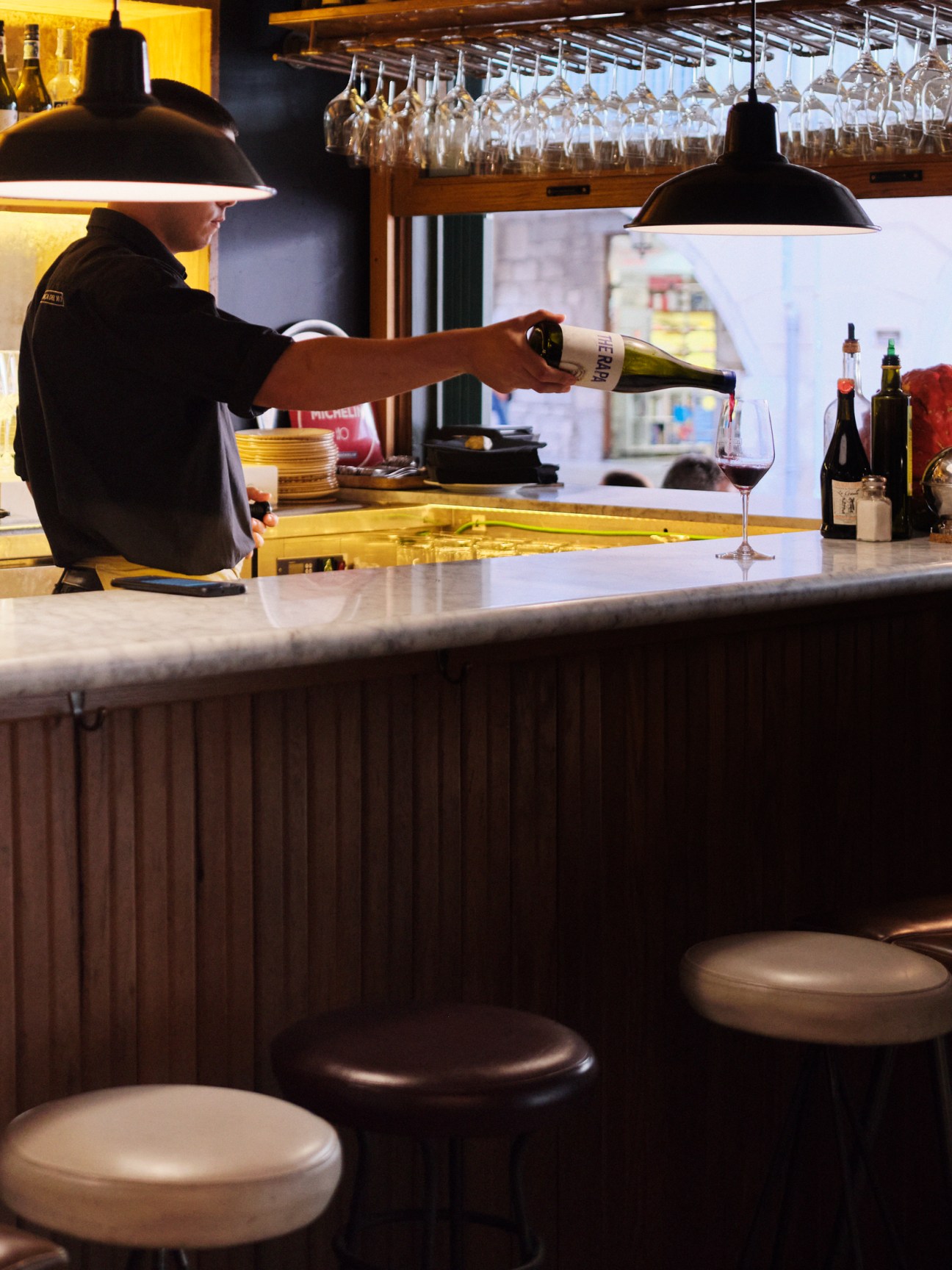 Bartender pouring wine at a bar counter with hanging glasses and black pendant lights.
