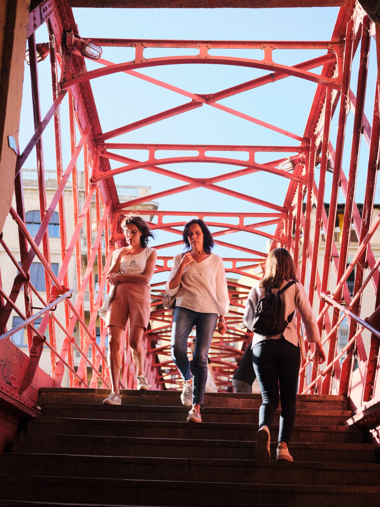 Three women jogging across a red metal bridge with geometric lattice design against blue sky.
