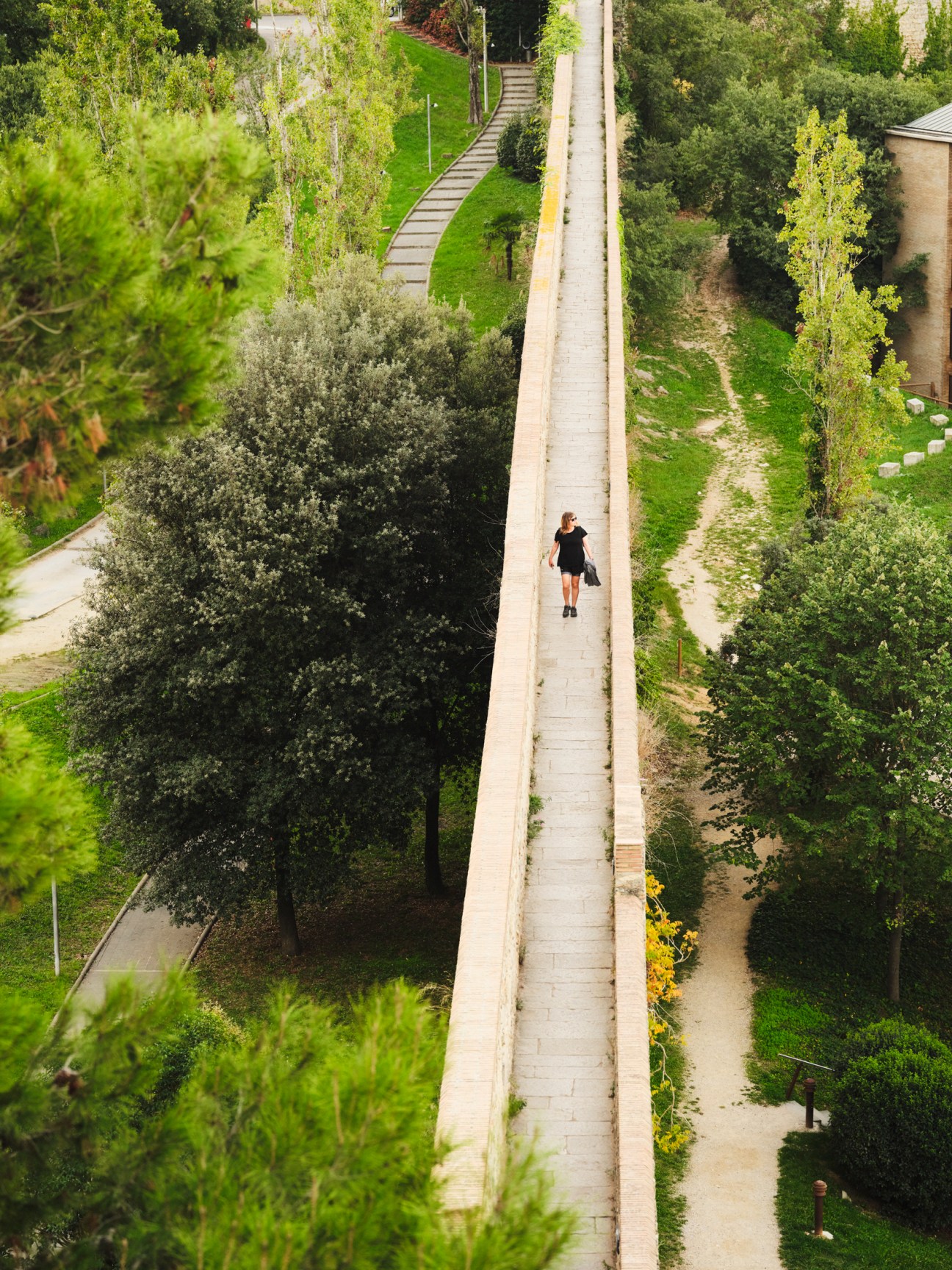 Woman walking along a narrow concrete pathway surrounded by lush green vegetation and trees.