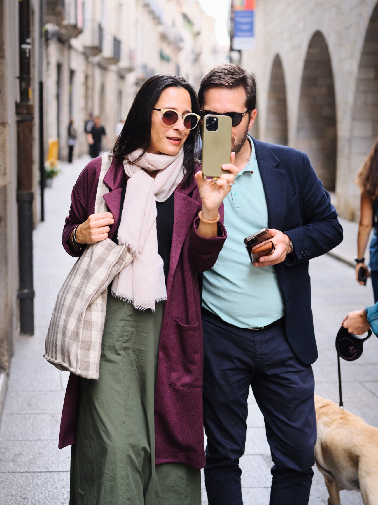 Two people walking through a narrow European street with historic stone buildings.