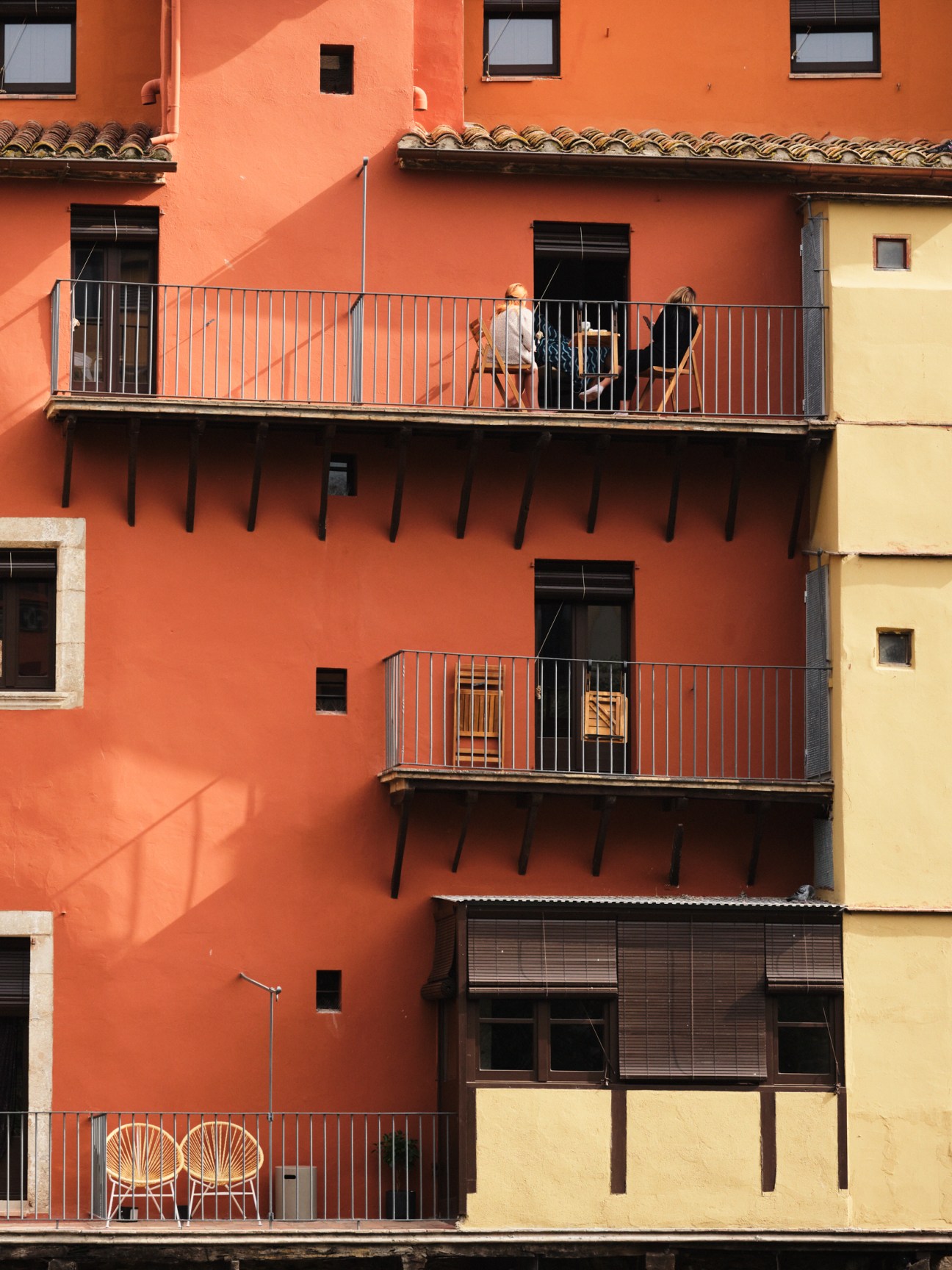 Bright orange residential building facade with metal balconies and wooden chairs in Girona, Spain.