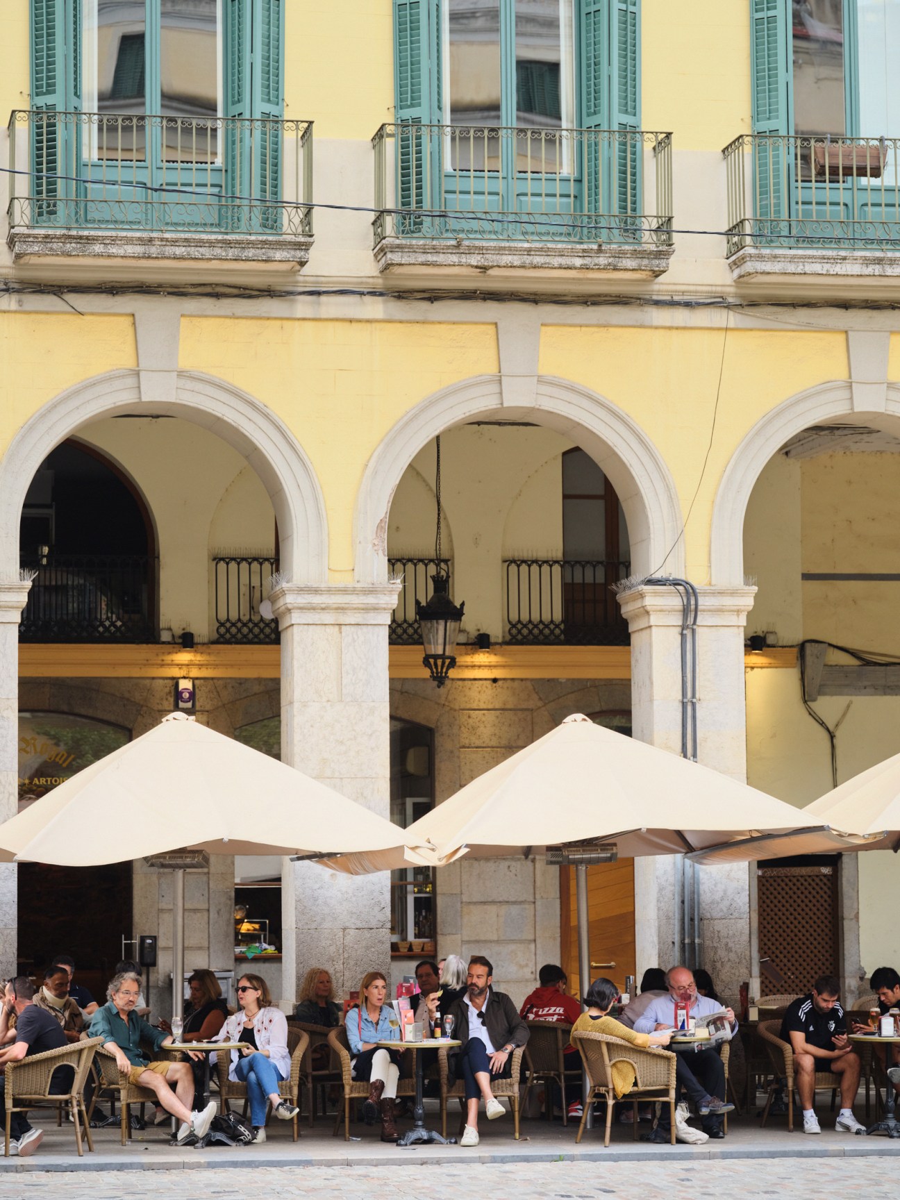 People sitting at outdoor café tables under white umbrellas in a yellow-walled plaza with arched colonnades in Girona.