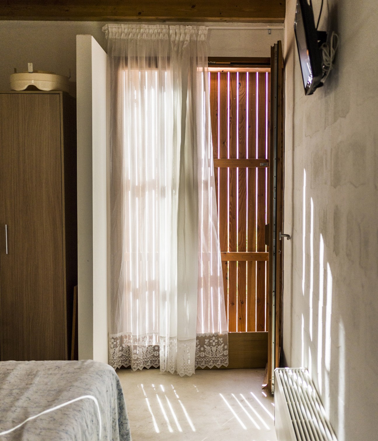 Bright bedroom with tall windows, sheer curtains, wooden wardrobe and decorative wooden door panels allowing natural light.