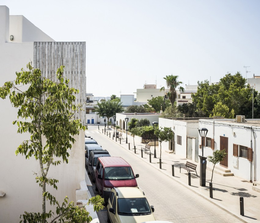 Peaceful pedestrian street in Formentera with white-walled houses, parked cars, and green vegetation under bright sunlight.