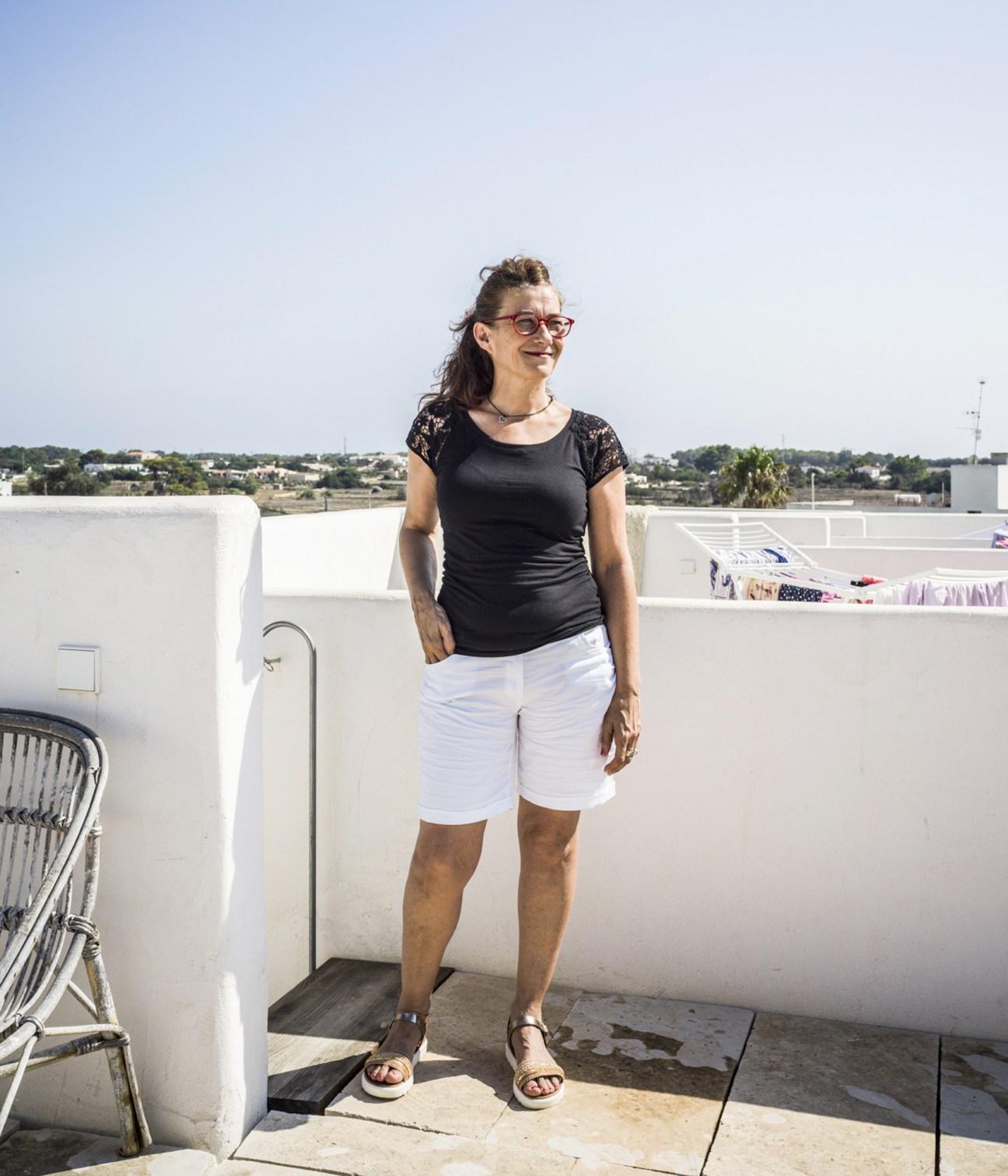 Woman wearing glasses and black lace-sleeved shirt standing on a rooftop terrace overlooking a coastal landscape.