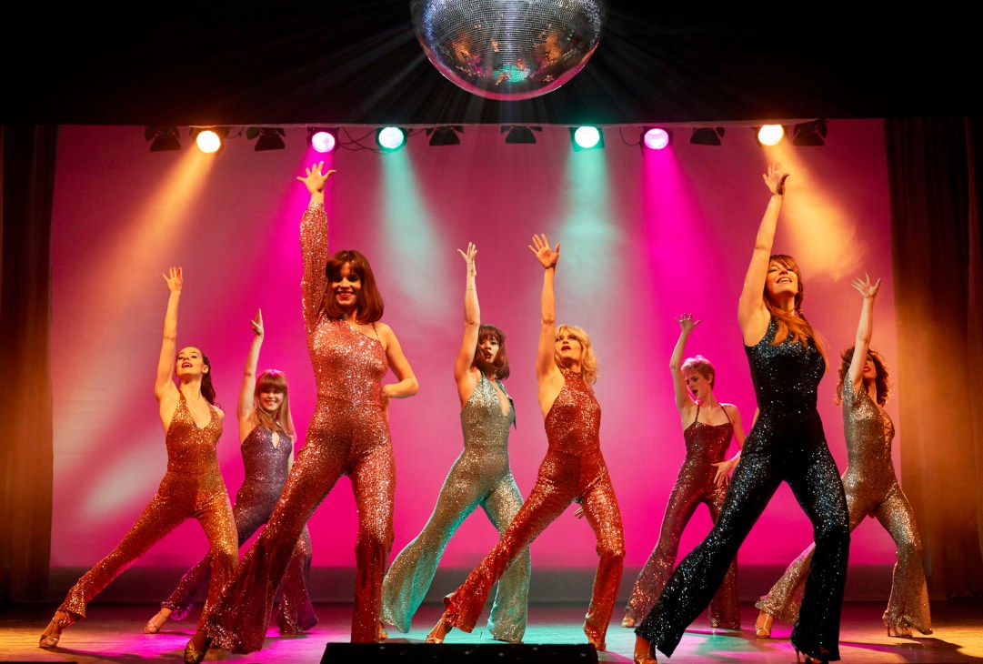 Group of female dancers in sequined costumes perform on a stage with pink lighting and a disco ball.