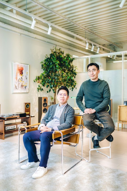 Two men pose in a modern studio space with exposed beams, vintage furniture, and potted plants.