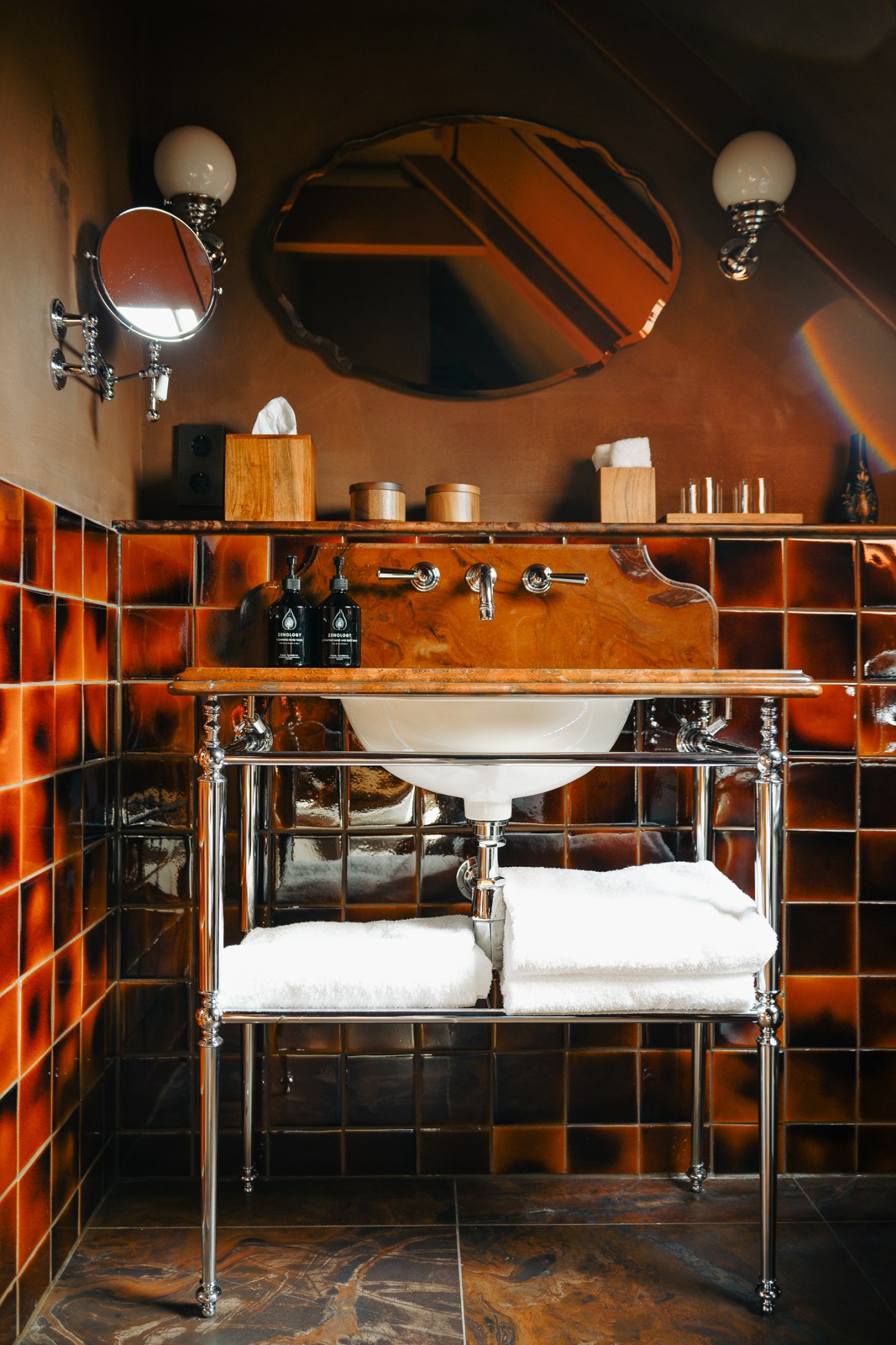 Luxury bathroom with orange-toned custom tiles, chrome washstand, oval mirror and vintage-style lighting fixtures