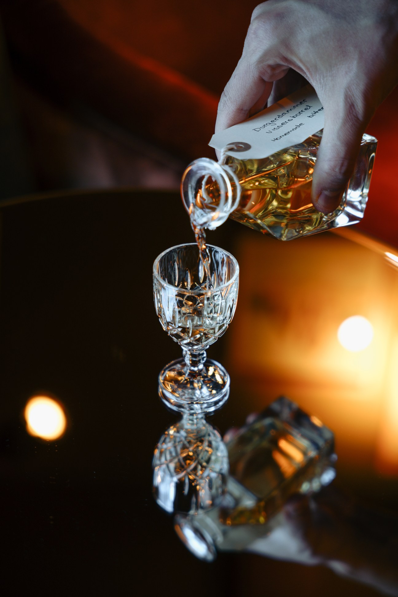 Hand pouring amber-colored spirit from crystal decanter into cut crystal glass on reflective surface