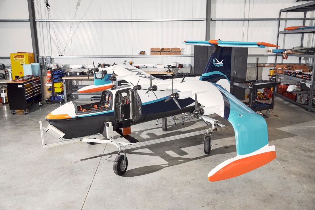 A sleek electric sea glider aircraft with blue, white and orange livery displayed in an industrial hangar