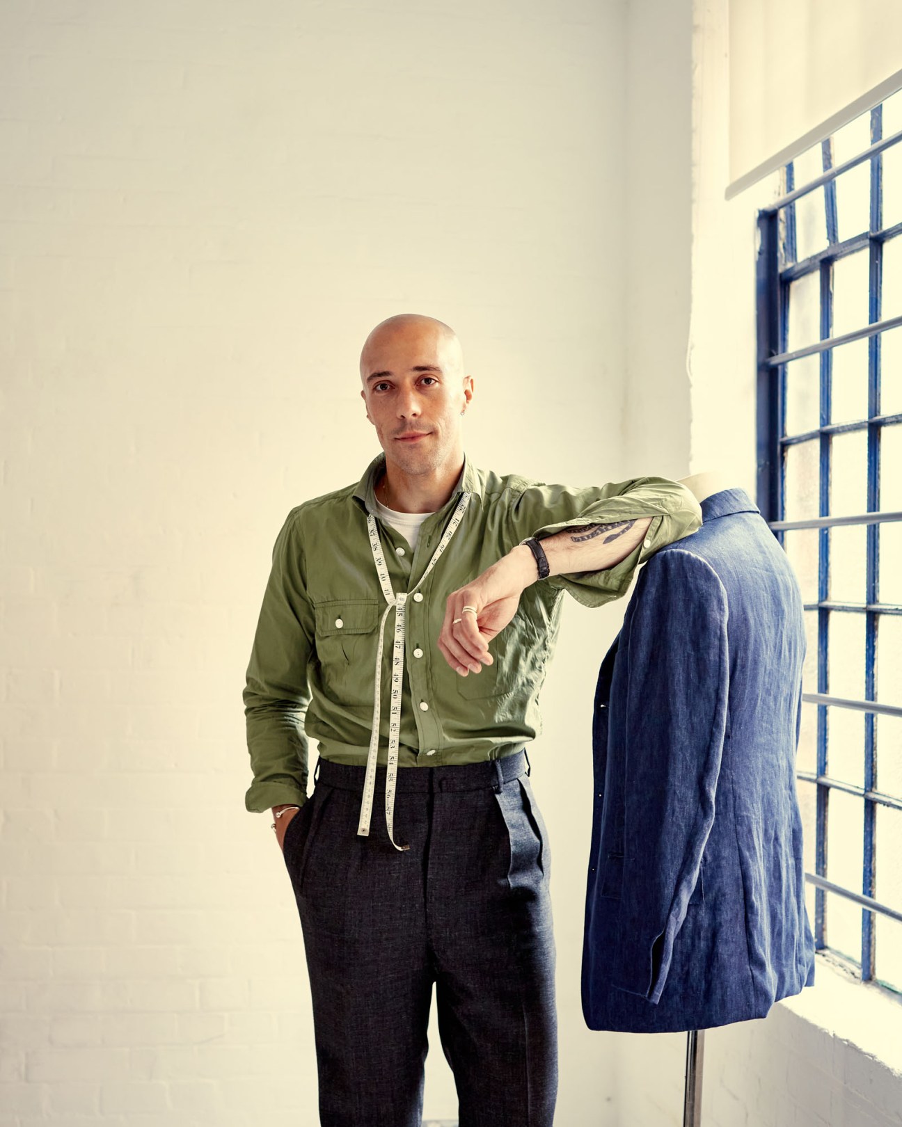 Fred Nieddu, a young tailor, wearing a green shirt and dark trousers stands in his London studio next to a blue jacket on a dress form.