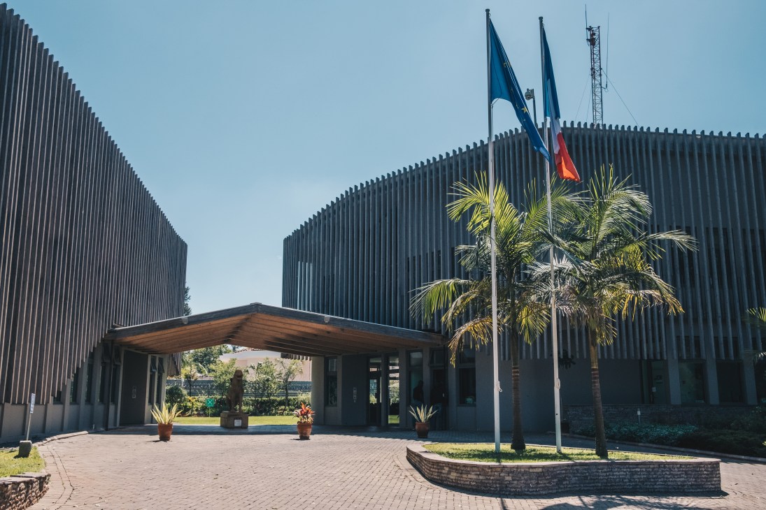 Modern embassy courtyard with curved wooden roof structure connecting two buildings, palm tree and French flag visible