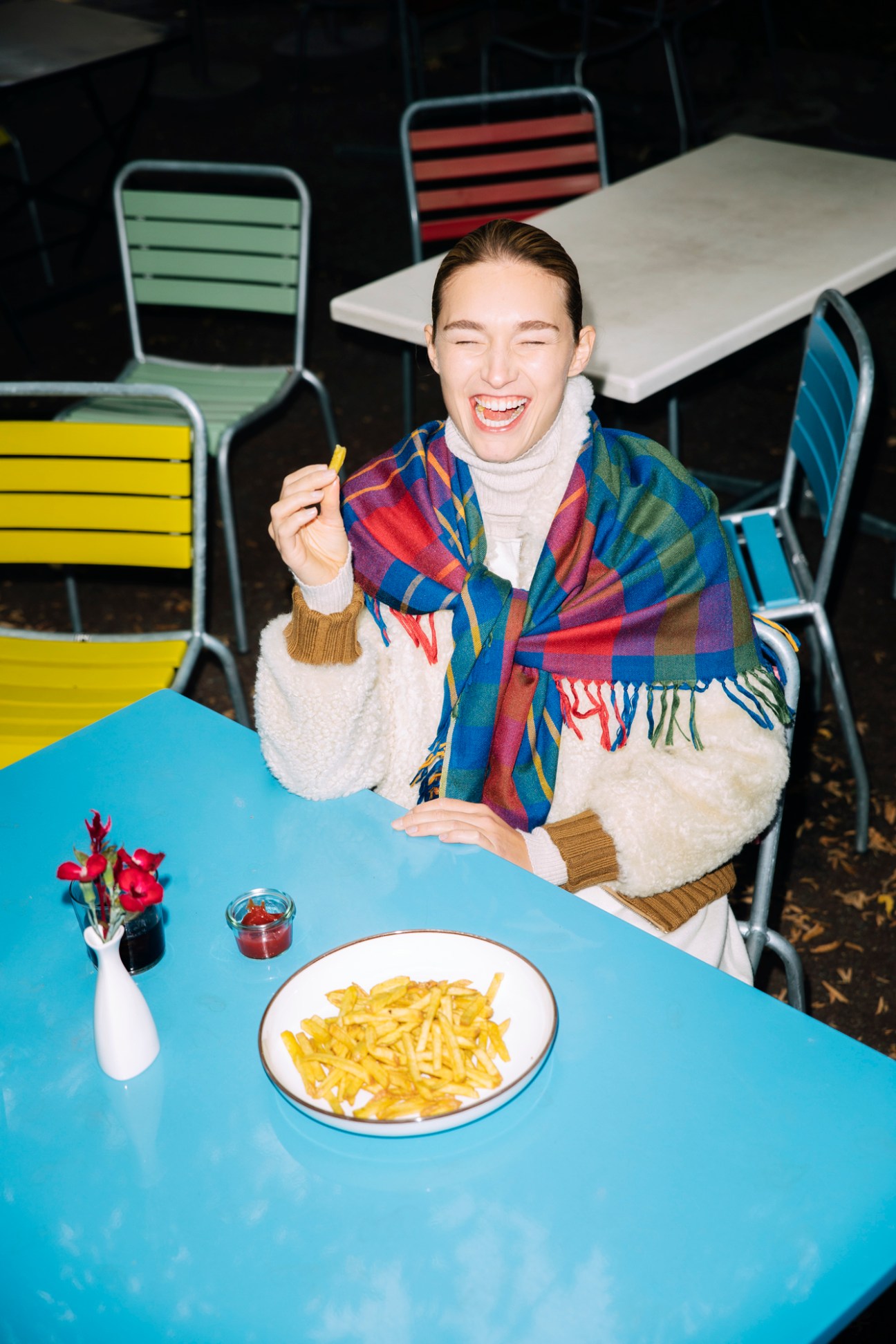 Model enjoying pasta at outdoor restaurant table wearing colorful plaid scarf and white turtleneck sweater