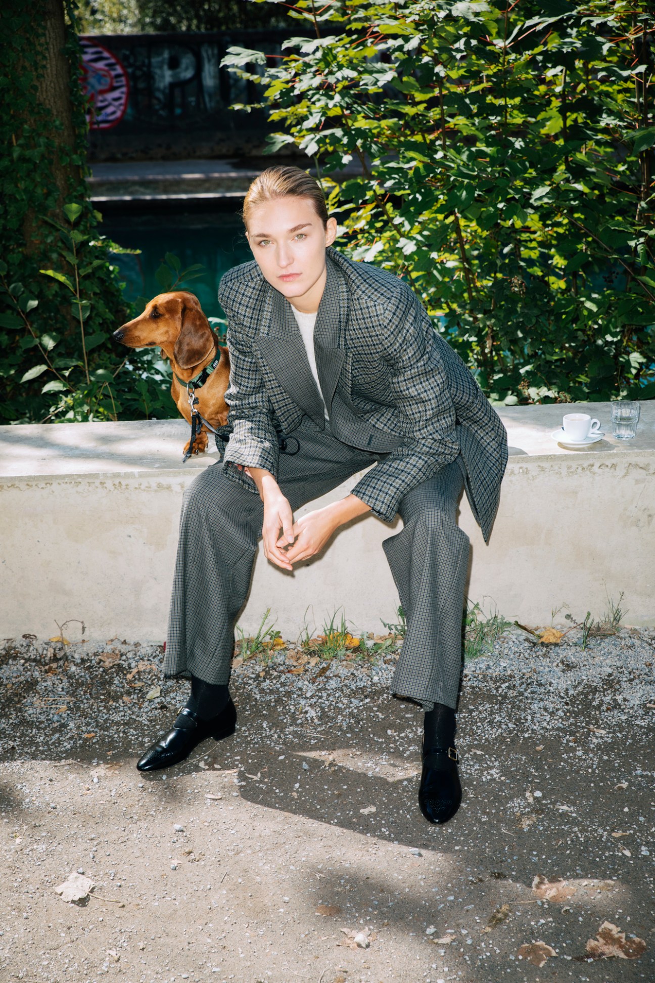 Model in grey checked suit sitting outdoors with dachshund dog, coffee cup visible on ledge