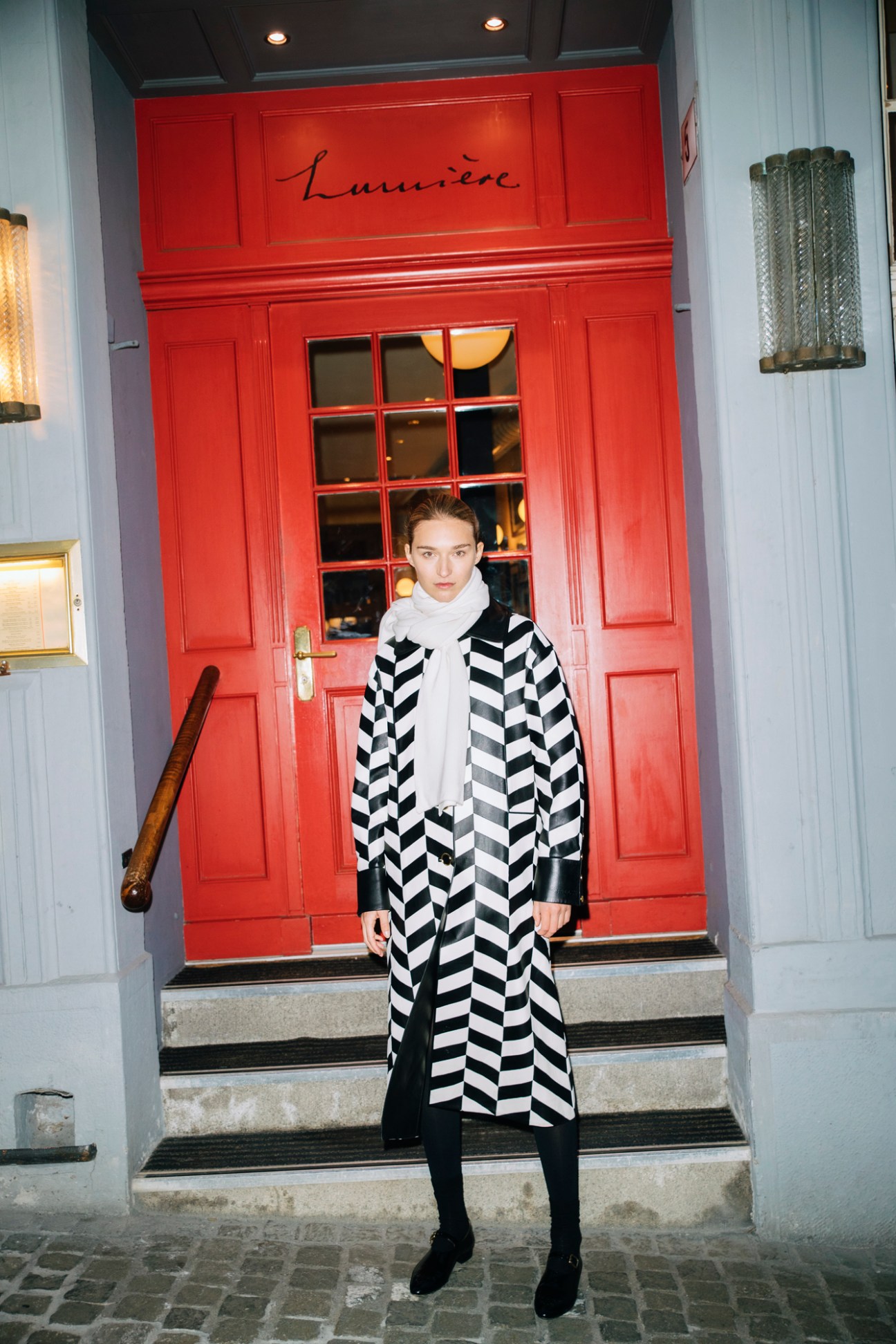 Model in black and white chevron coat with white turtleneck standing on steps outside restaurant Lumiere