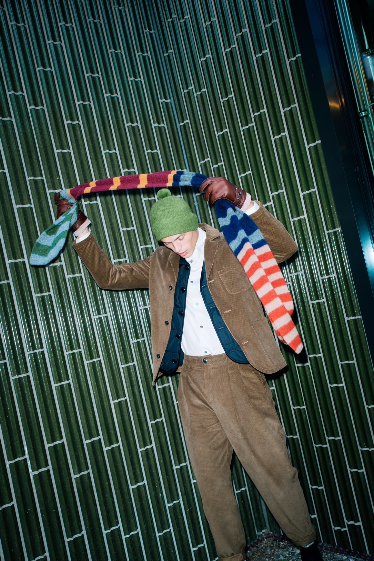 Model in brown suit and green beanie holding colorful striped scarf above head against bamboo backdrop