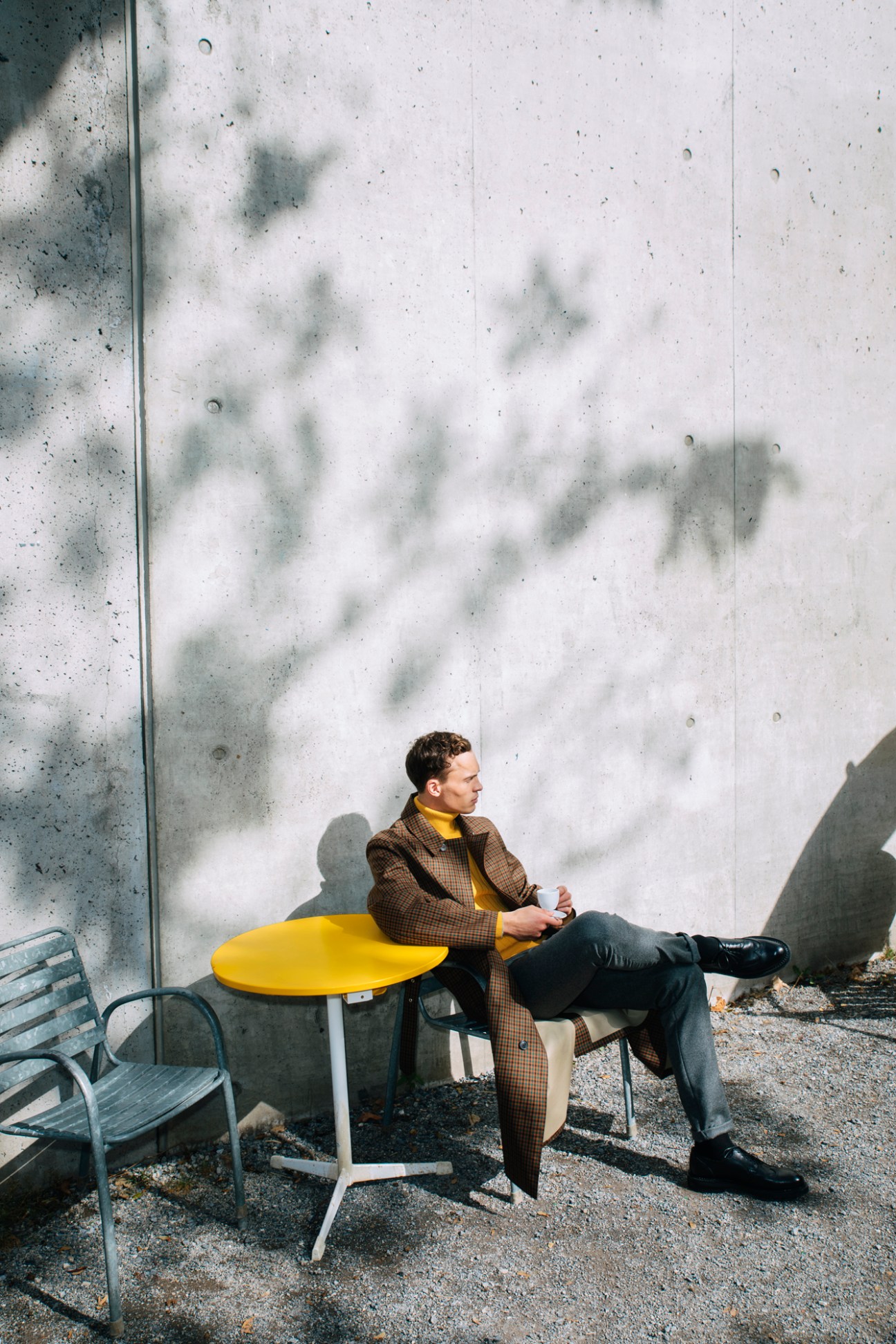Man in brown plaid coat and yellow sweater sitting at yellow table outside concrete wall