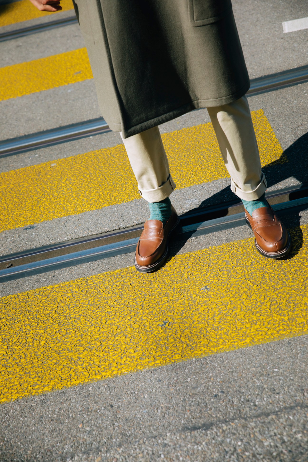 Person's legs in beige trousers, green socks and brown leather loafers standing on yellow striped pavement
