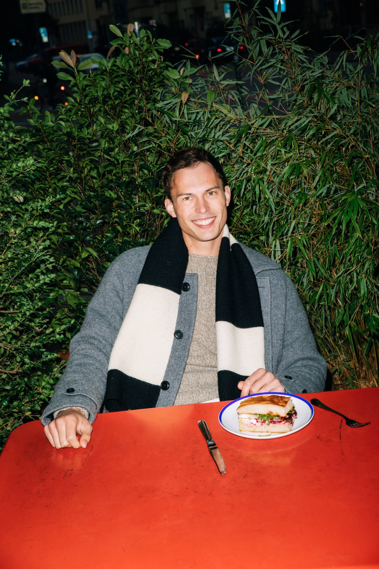 Model wearing Aspesi coat with Howlin' jumper and Margaret Howell scarf dining outdoors at red table