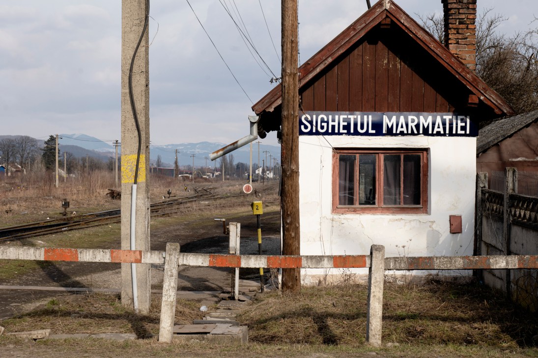 A small railway station building with 'SIGHETUL MARMATIEI' sign and train tracks in rural mountainous setting