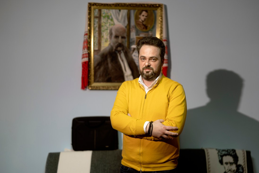 Nicolae-Miroslav Petretchi in yellow sweater standing in office with framed portrait and flags behind him