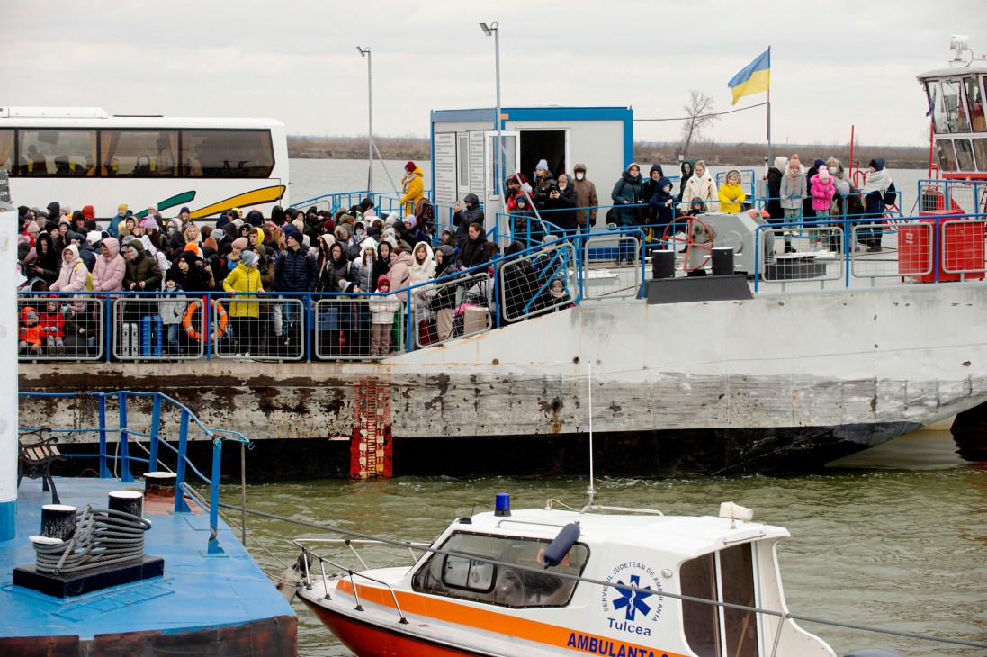 Crowds of refugees with luggage and belongings gathered on a ferry dock with Ukrainian flag visible
